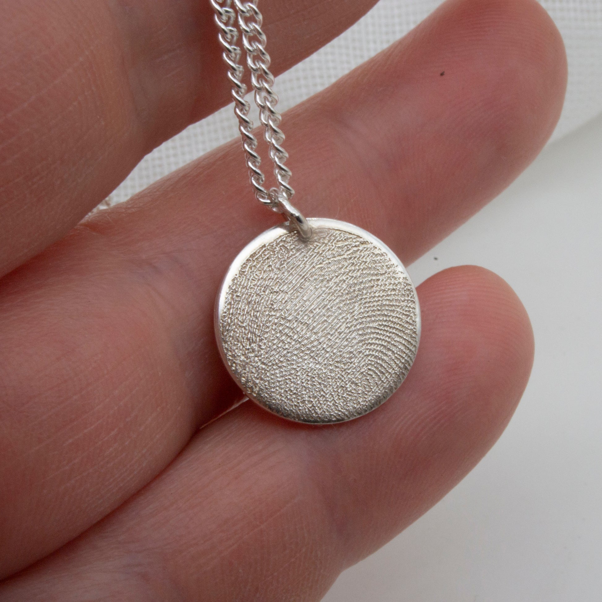 Silver textured pendant on a chain held between fingers against a white background